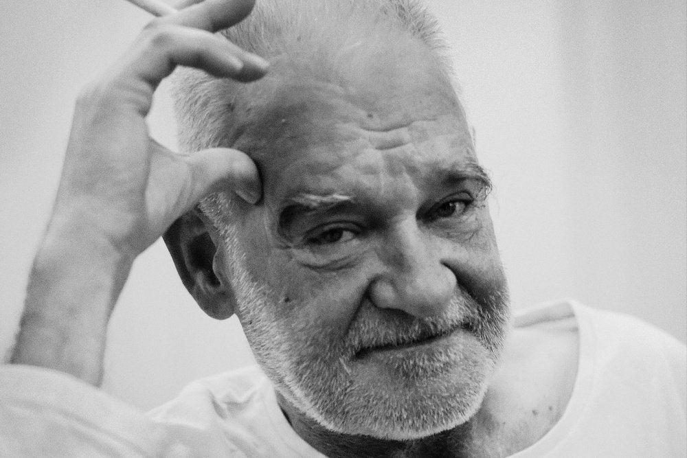 Black-and-white close-up of an older, gray-bearded man in a white shirt resting his hand against his temple.