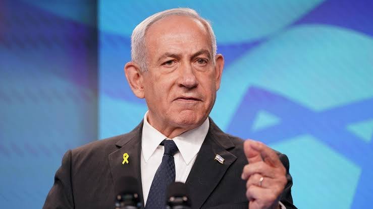 An older man with white hair, wearing a dark suit and blue tie, speaks at a podium with a yellow ribbon pin, against a blue backdrop featuring a Star of David.
