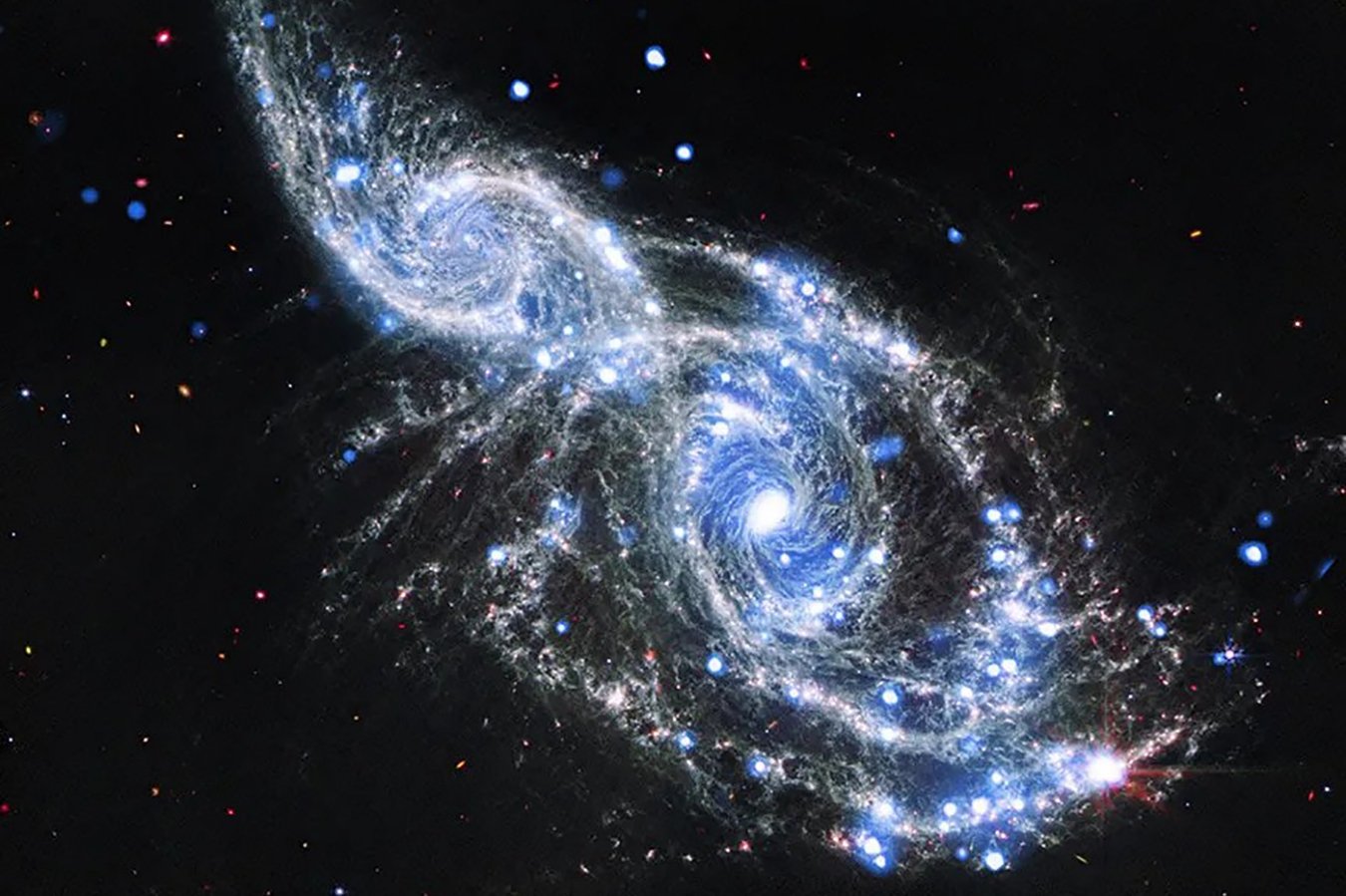 Two interacting spiral galaxies with bright blue star-forming regions and glowing cores appear to merge against a dark background dotted with distant stars.