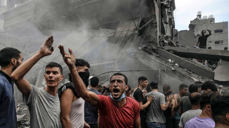 A crowd of men shout and gesture amid heavy smoke in front of a severely damaged, collapsed concrete building.