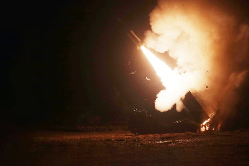 A missile launcher fires a projectile at night, producing a bright orange flare and large plume of smoke over a dark field.
