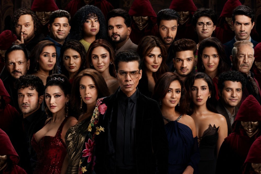 A group of approximately twenty people pose together against a dark background, with a man in a black suit centered and masked figures in red hoods visible behind them.