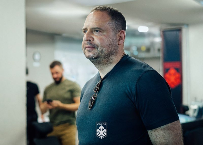 A bearded man in a dark blue t-shirt with sunglasses tucked into the collar stands indoors, while another man in an olive shirt looks at a phone in the background.