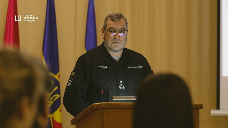 A bearded man in a dark uniform stands at a wooden podium, facing an audience, with Ukrainian flags visible behind him.