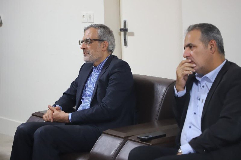 Two men in dark suits sit on brown leather chairs against a white wall, one with clasped hands and the other resting his hand near his face.