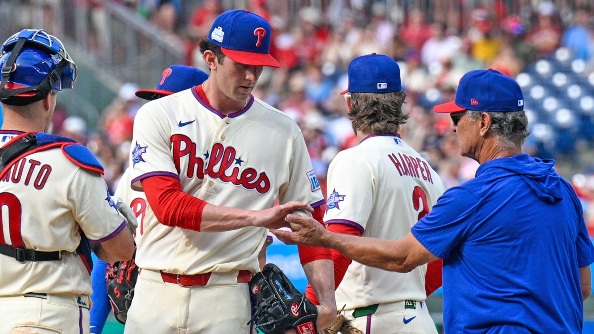 Phillies pitcher hands the baseball to a coach on the mound during a mound visit, with catcher and "HARPER" visible in the background.