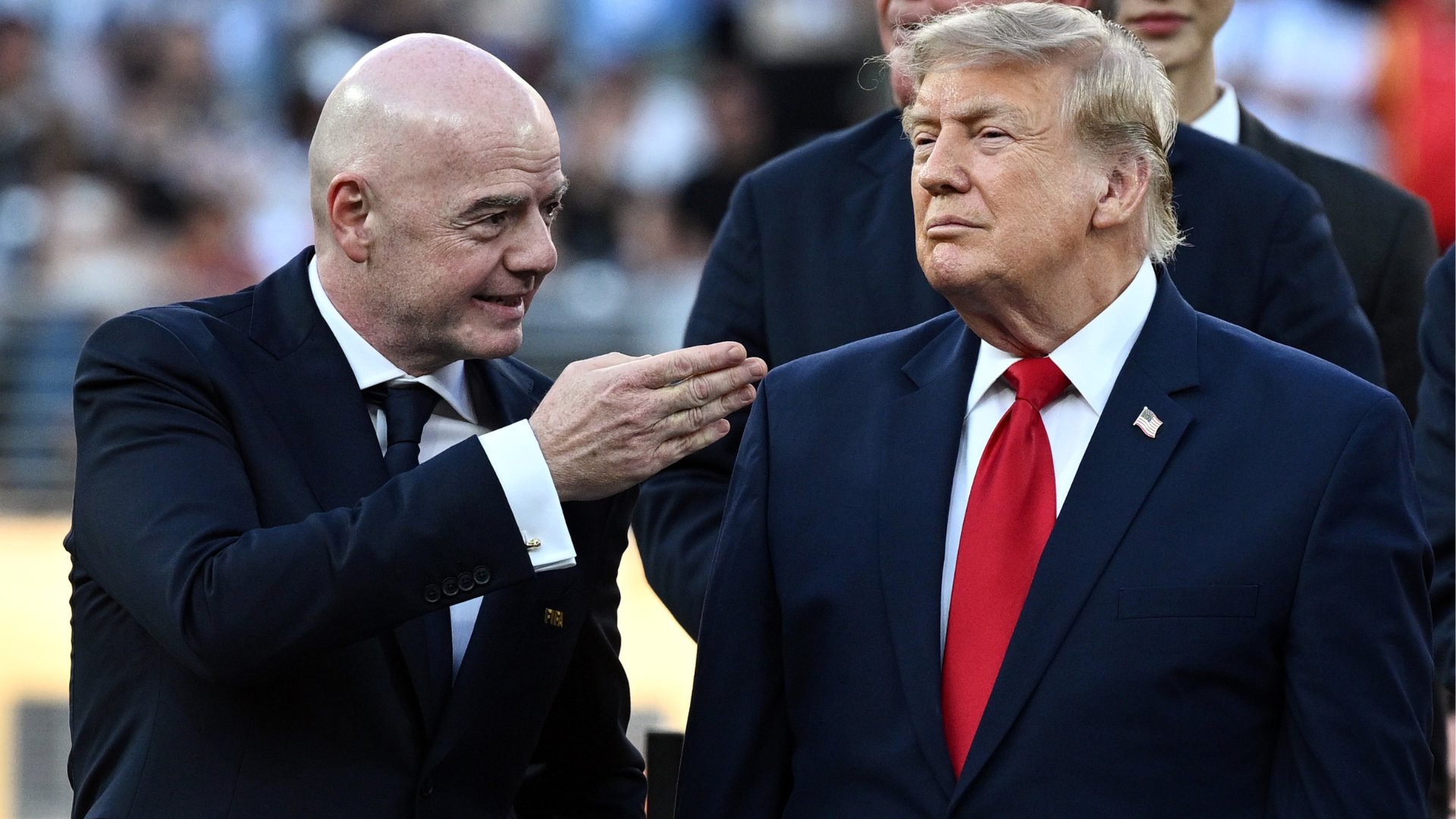 A bald man in a dark suit gestures toward a blonde man in a navy suit and red tie, who wears an American flag lapel pin, amid a blurred crowd.