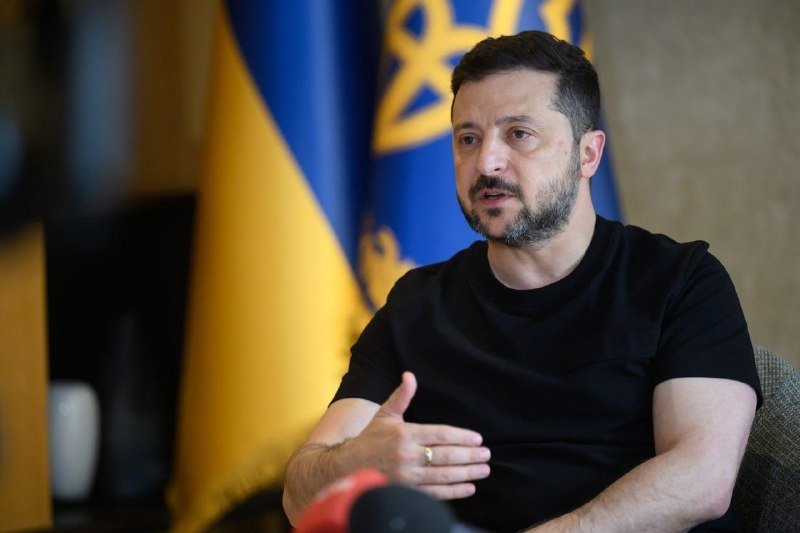 A bearded man in a black t-shirt speaks while gesturing with his hand, seated in front of the blue and yellow Ukrainian flag.