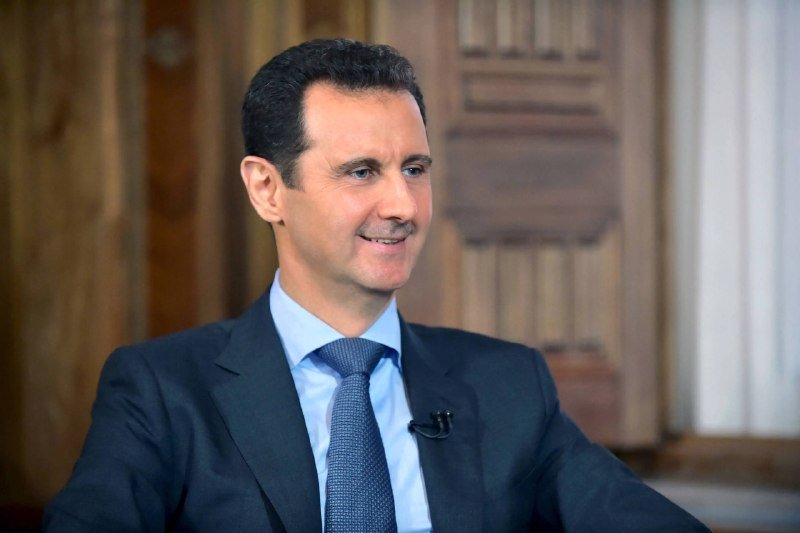 A smiling man in a dark suit, light blue shirt, and patterned tie speaks during an interview, with a small lapel microphone clipped to his jacket.