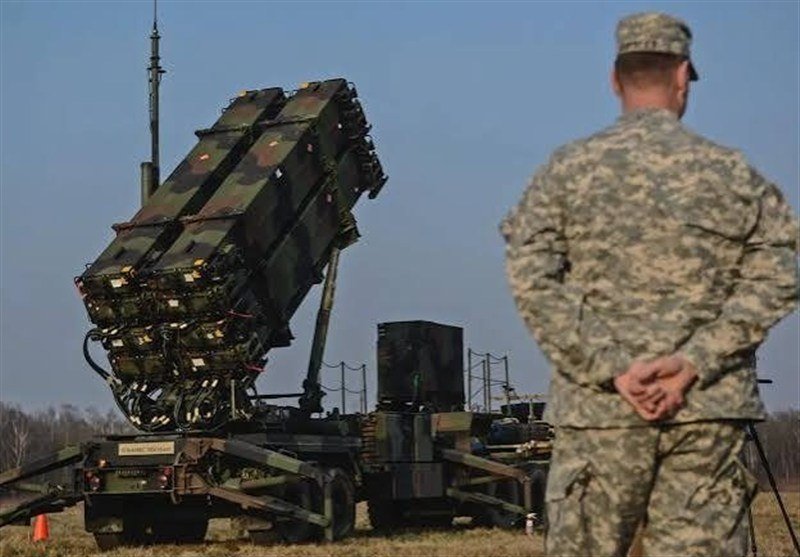 A soldier in camouflage uniform stands with hands clasped behind his back, facing a raised mobile missile launcher system positioned in a grassy field under a clear sky.