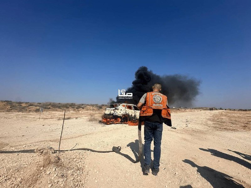 A man in an orange vest stands in a desert landscape, watching a burning vehicle billowing thick black smoke into a clear blue sky.