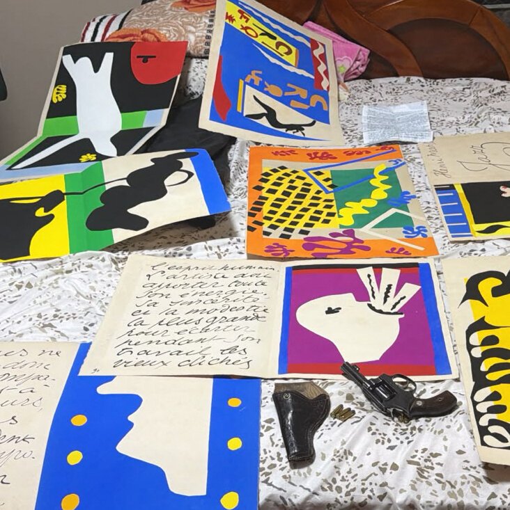 Colorful abstract paintings and handwritten notes, including one with French text, are spread across a floral-patterned tablecloth alongside a small black gun-shaped object.
