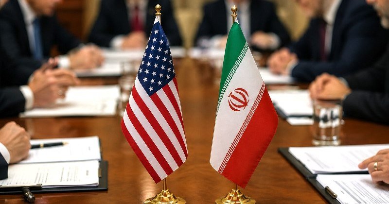 Small U.S. and Iranian flags stand side by side on a conference table as suited officials sit with documents before them.