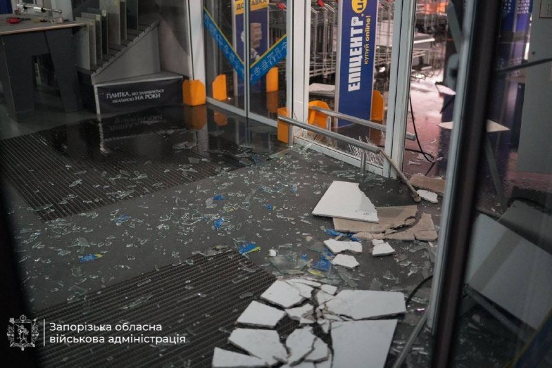Shattered glass and broken tiles litter the floor of a damaged storefront with Ukrainian signage, including a "Zaporizhzhia Regional Military Administration" emblem in the corner.