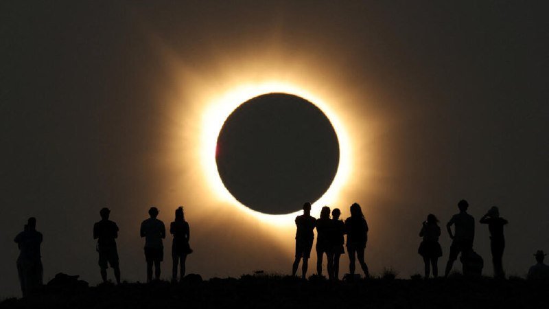 Silhouetted people stand on a ridge watching a total solar eclipse, the darkened moon surrounded by a glowing ring of light against a darkened sky.