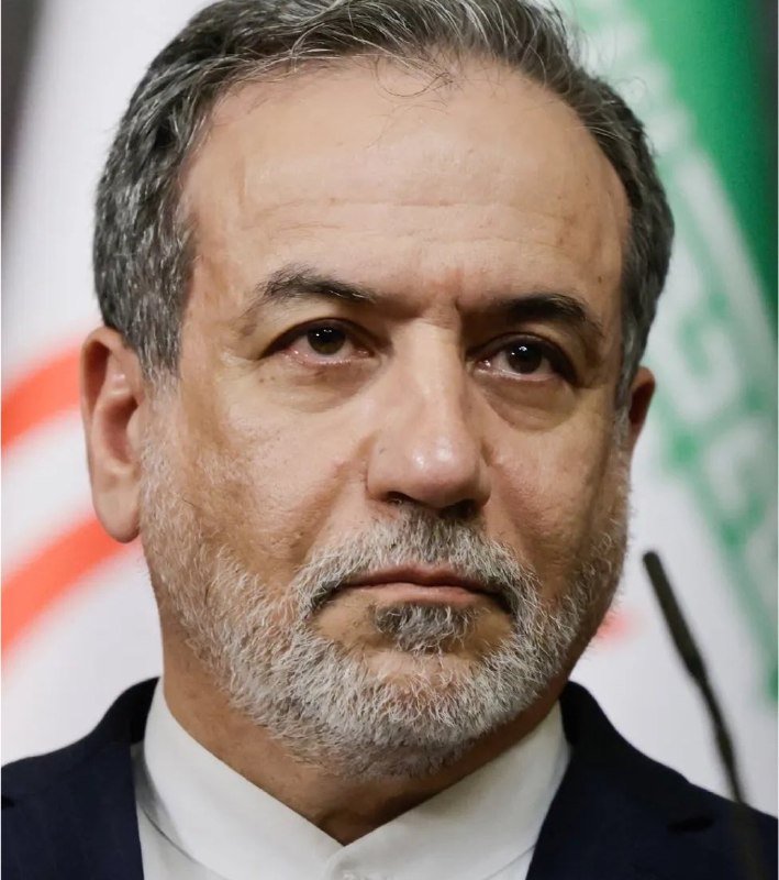 A close-up photo shows a middle-aged man with gray hair and beard, wearing a dark suit jacket and white collared shirt, positioned in front of a partially visible green and white flag.