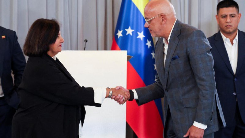 A woman in a black blazer and a bald man in a gray checkered suit shake hands in front of a Venezuelan flag, while a man in a dark suit stands behind.