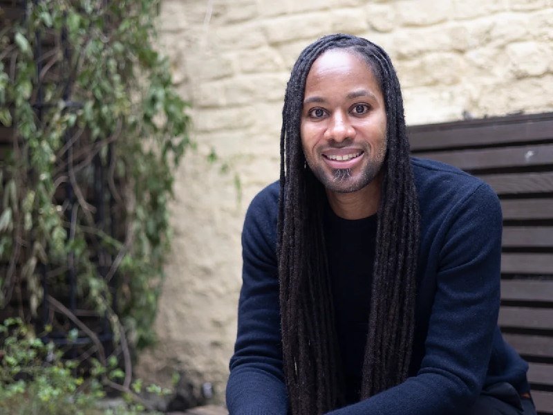 A man with long dreadlocks and a goatee smiles while seated outdoors against a stone wall with green vines visible to the side.