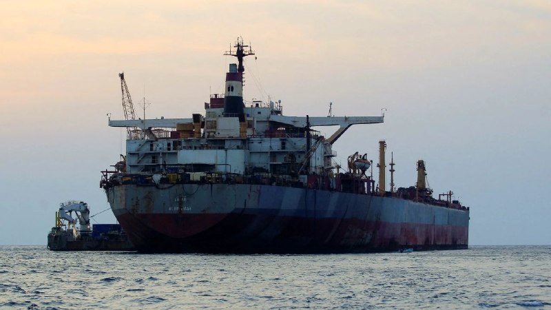 A large rusted tanker ship floats on calm open water, accompanied by a small tugboat alongside, under a pale hazy sky at sunset.