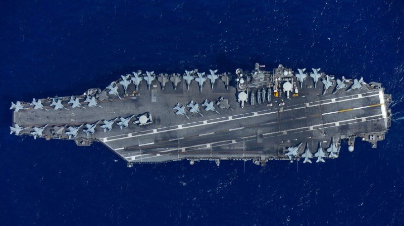 An aerial view shows a large aircraft carrier loaded with numerous military jets sailing on dark blue open water.