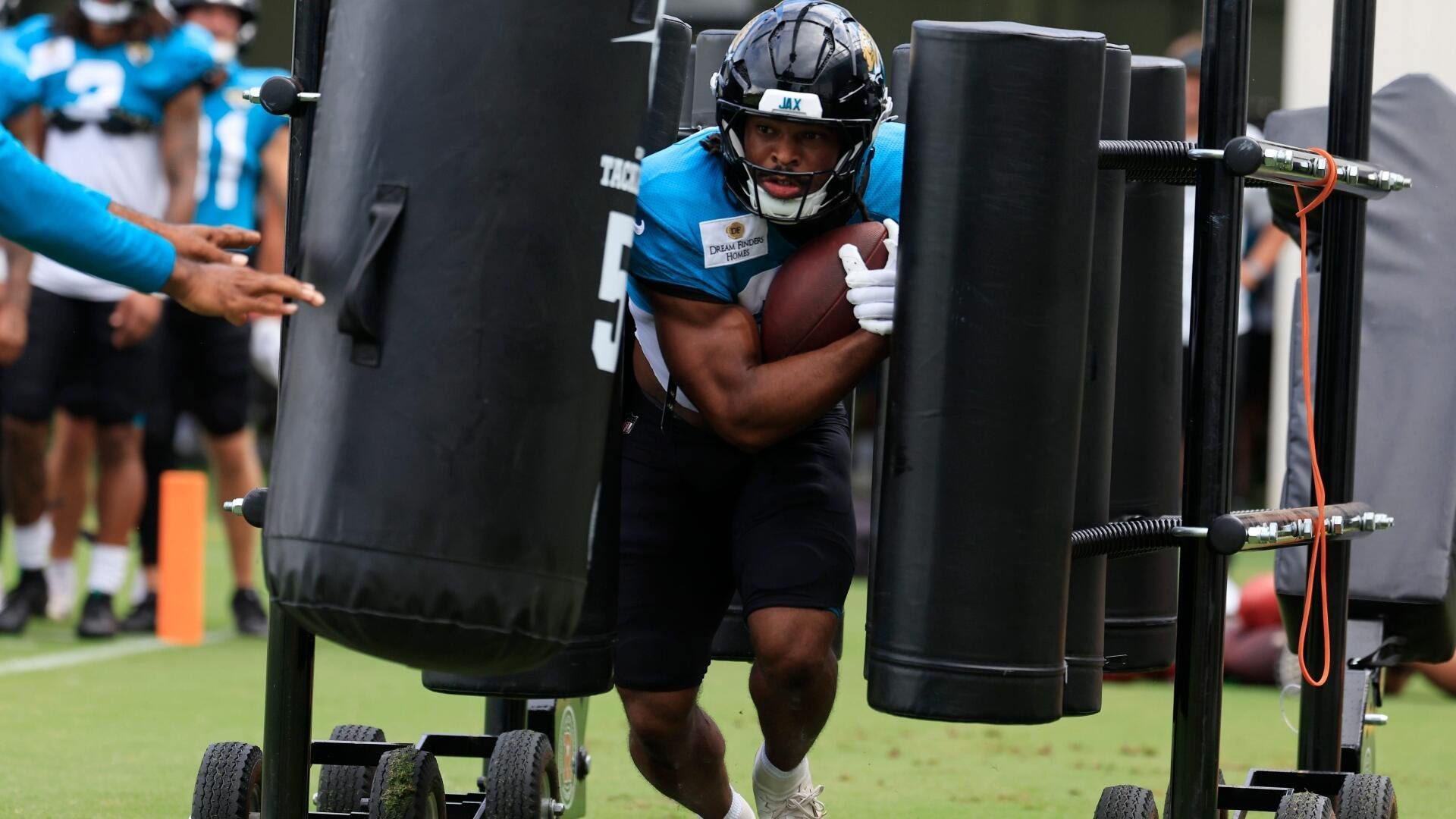 A football player in a teal jersey and helmet carries a football while running between padded blocking sleds during an outdoor practice.