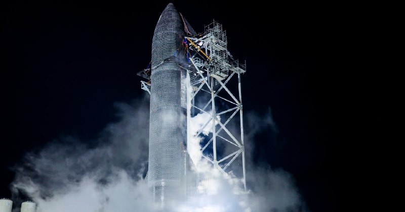 A large, silver, bullet-shaped rocket stands on a launch pad at night, surrounded by metal scaffolding with white smoke and vapor venting from its base.