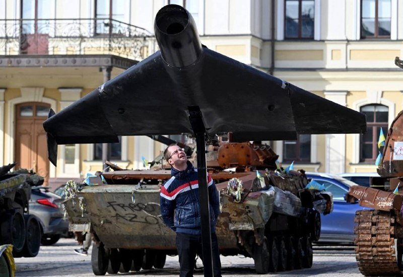 A man in a blue, white, and red jacket stands in front of a display of damaged, burnt military tanks on a city street, with a large dark aircraft component positioned above him.