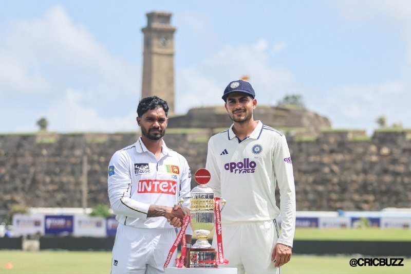 Two cricket players in white uniforms pose alongside a silver trophy in front of an ancient stone tower and fortress on a sunny day.