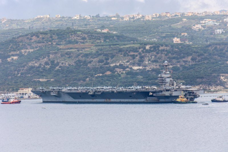 A large gray aircraft carrier with multiple fighter jets on its deck floats in calm waters, accompanied by smaller tugboats, with a hillside dotted with houses in the background.