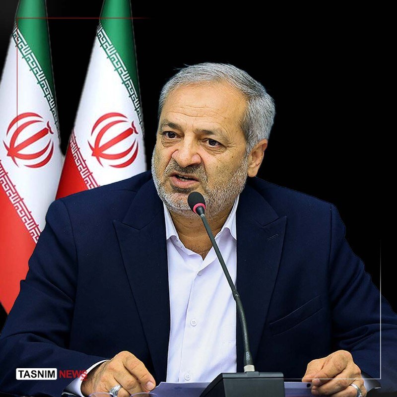 A gray-bearded man in a dark suit and white shirt speaks into a microphone, with two Iranian flags visible in the background.