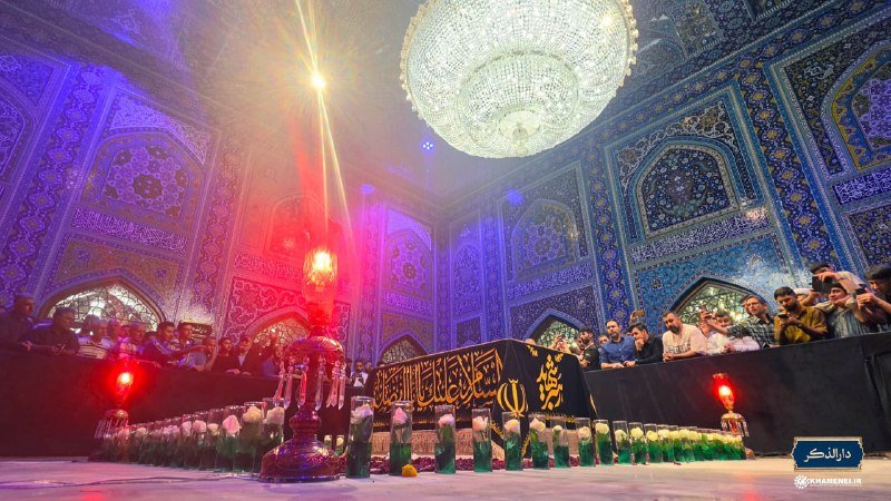 A crowd gathers inside an ornately tiled shrine around a black-draped tomb adorned with Arabic calligraphy, illuminated by a crystal chandelier and lit candles.