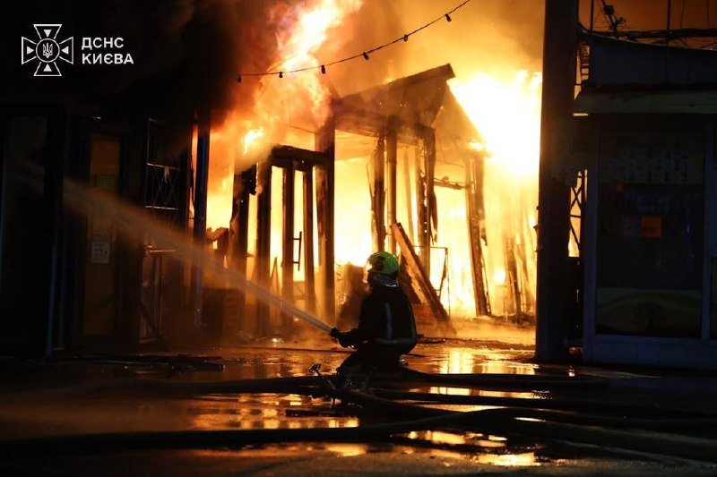 A firefighter in a helmet sprays a water hose at a large, intense fire engulfing a building's structure at night.