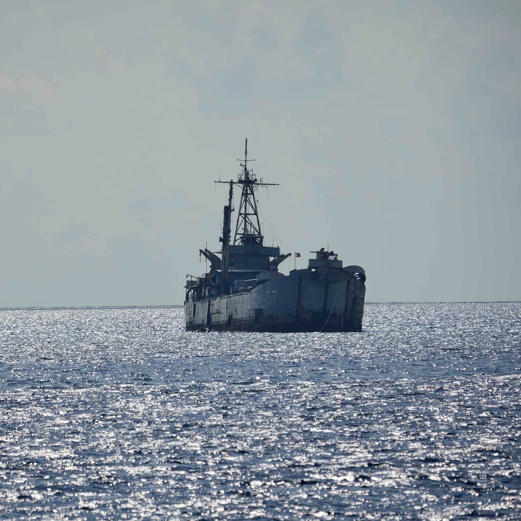 A rusted, derelict naval vessel with a tall mast sits grounded in shallow, sunlit ocean water under a hazy sky.