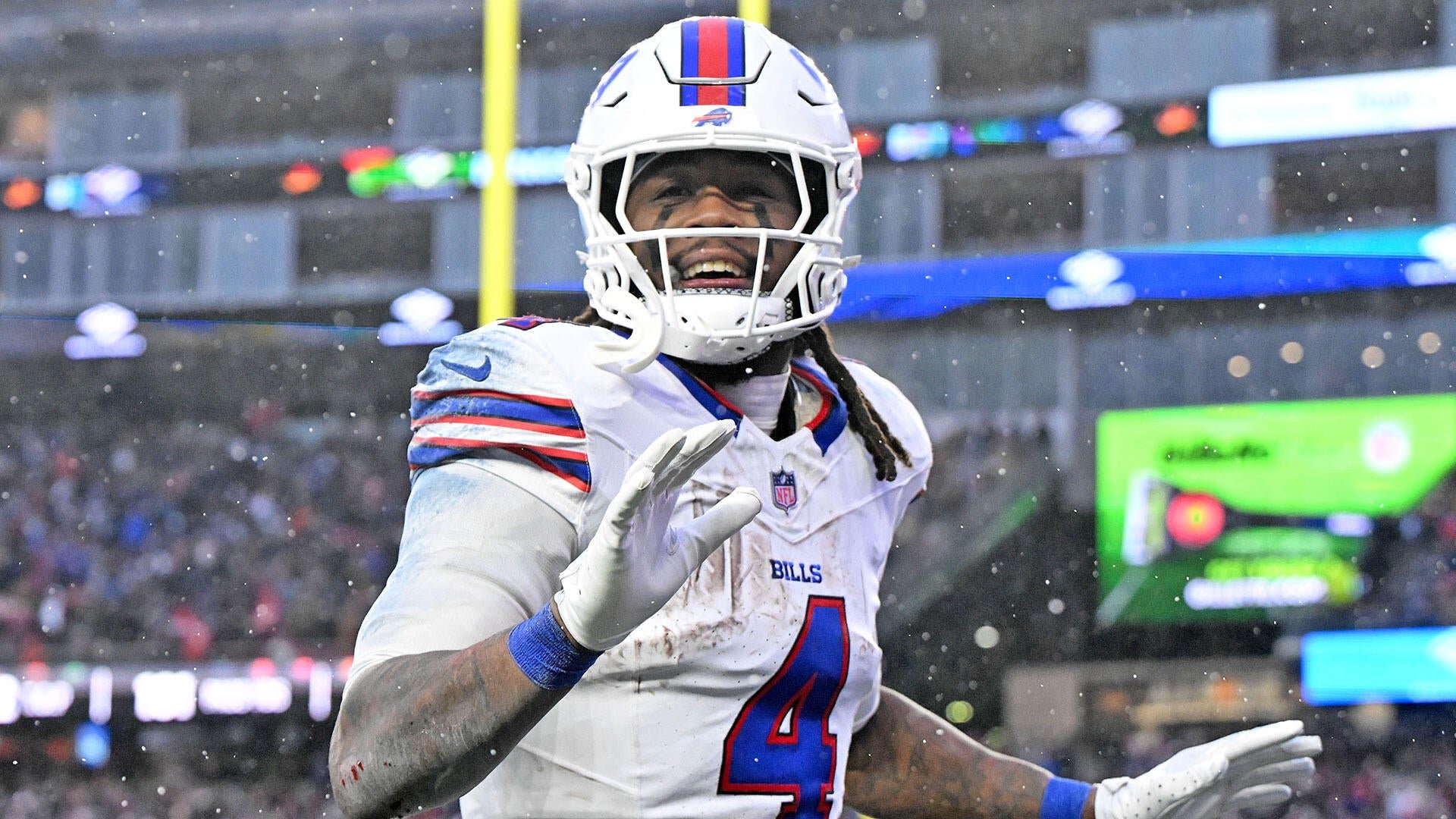 A football player wearing a white Bills jersey with the number 4 smiles and raises his gloved hands while snow falls around him in a stadium.