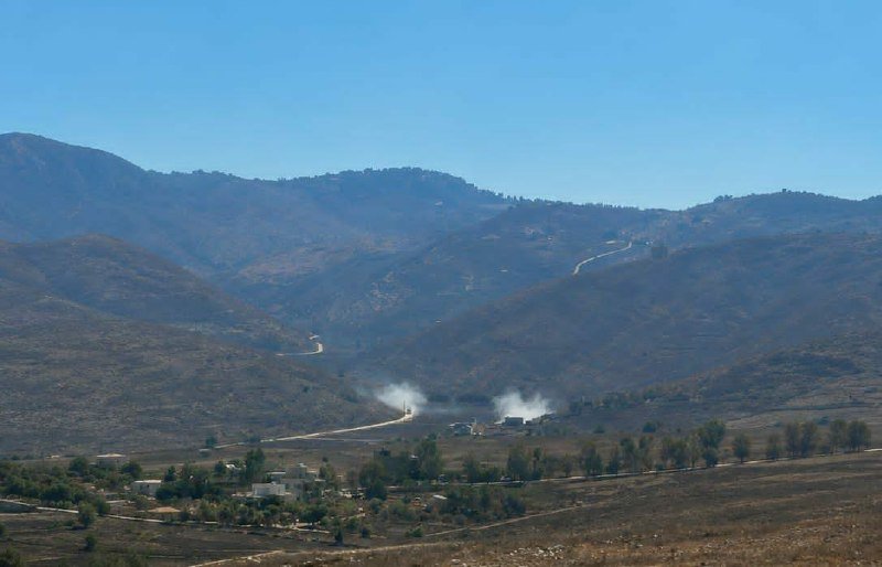 Smoke rises from vehicles traveling along a dirt road through a mountainous, arid landscape with scattered small buildings and trees in the foreground.
