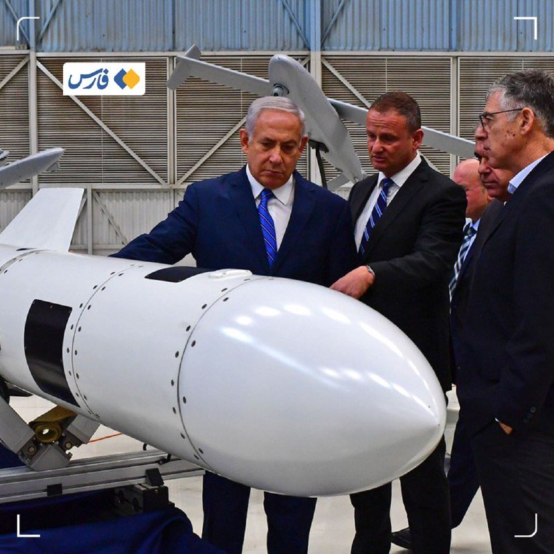 A man in a blue suit and tie stands beside a large white missile-shaped object alongside other suited men inside a hangar-like facility.