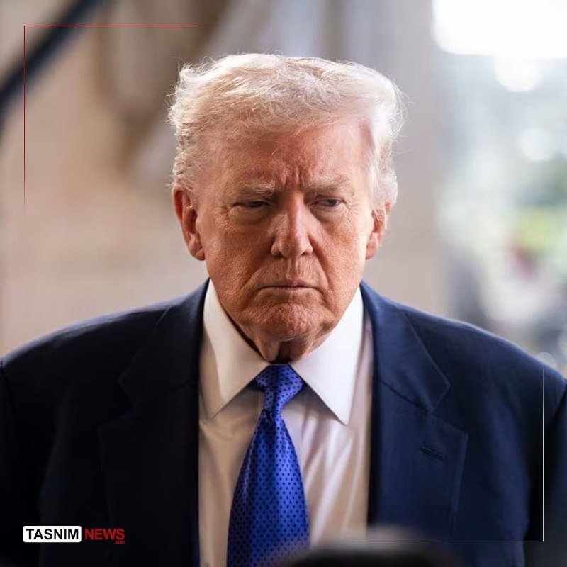 An older man with light-colored hair, wearing a dark suit, white shirt, and blue tie, looks downward with a serious expression.