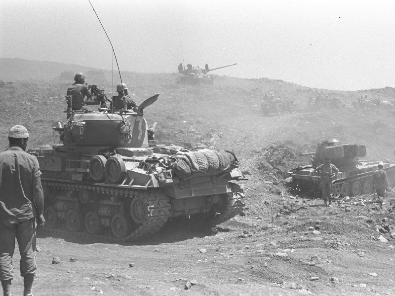 Black-and-white photo shows soldiers and tanks advancing across a dusty, rocky hillside terrain.