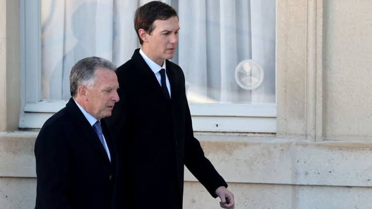 Two men in dark suits and ties walk past a building with large windows.