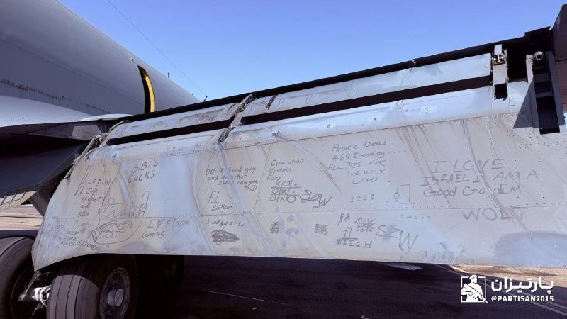 A heavily scrawled aircraft fuselage covered in handwritten graffiti messages sits on tarmac next to a truck, under clear blue skies.