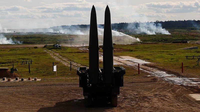 Two missiles stand vertically on a mobile launcher in an open field, with smoke plumes visible in the distant background under a partly cloudy sky.