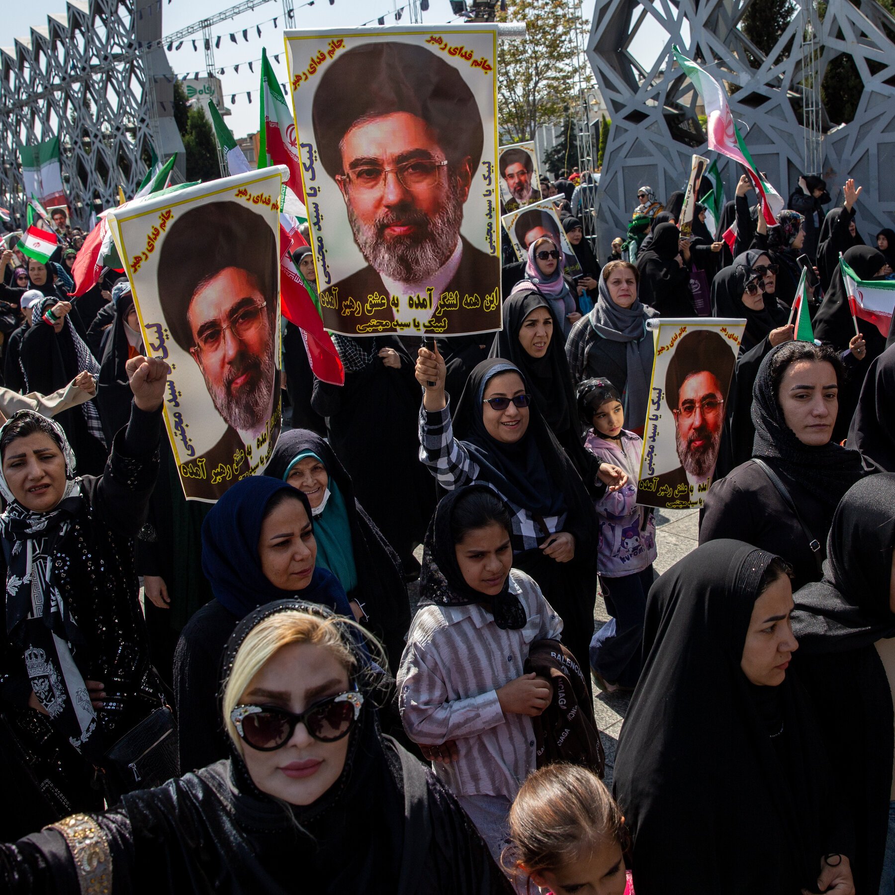 A crowd of women and children in black headscarves march outdoors, holding Iranian flags and posters displaying the same bearded portrait of a man wearing glasses and a turban.