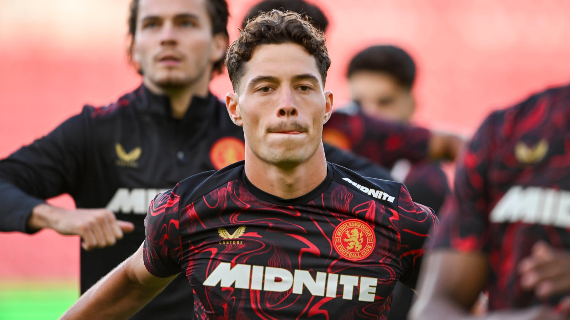 Middlesbrough football players wearing red and black striped jerseys with the "MIDNITE" sponsor logo perform warm-up exercises on a pitch.