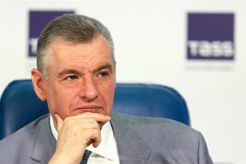 A grey-haired man in a grey suit and patterned tie sits with his hand resting against his chin, looking off to the side against a blurred background with purple TBS logos.