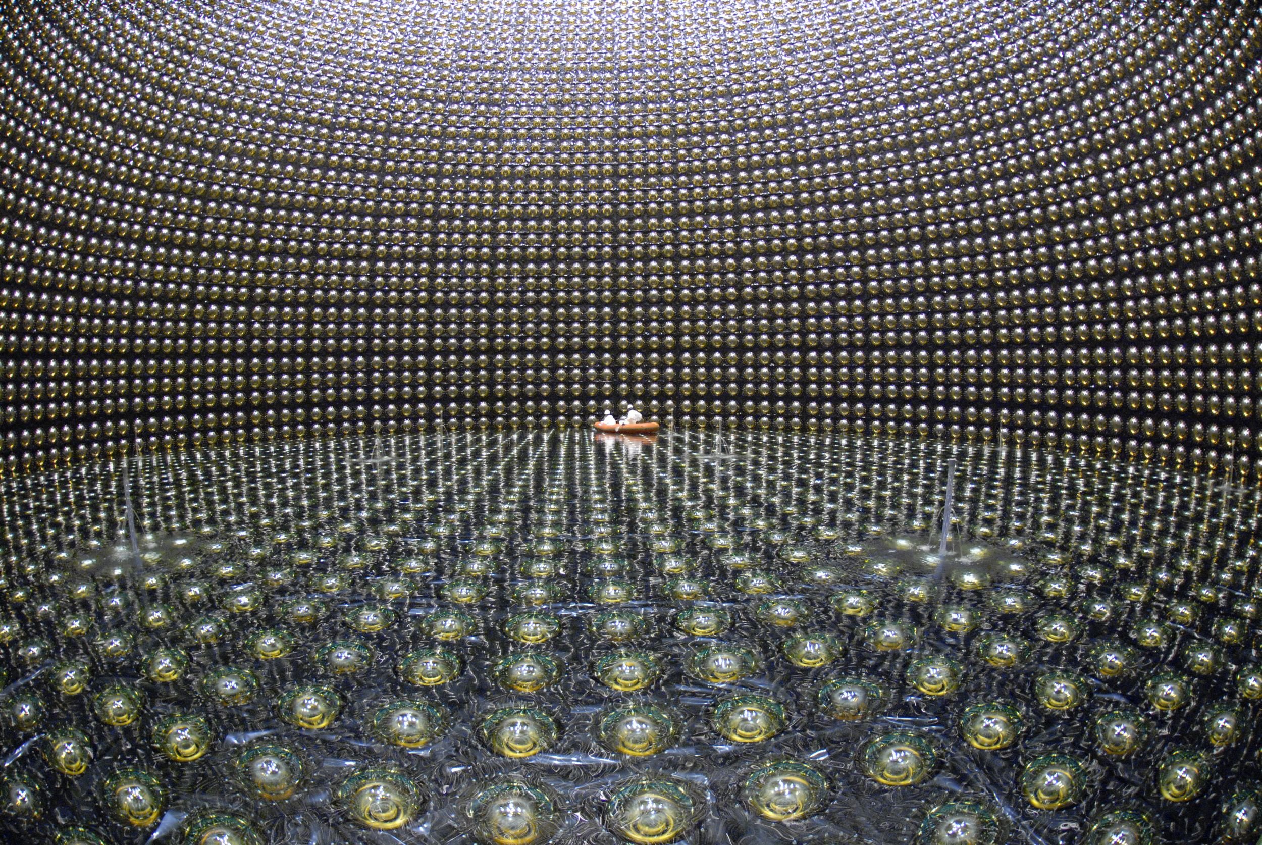Two small figures in white helmets float in an orange raft on water surrounded by thousands of spherical sensors lining a massive underground detector's walls.