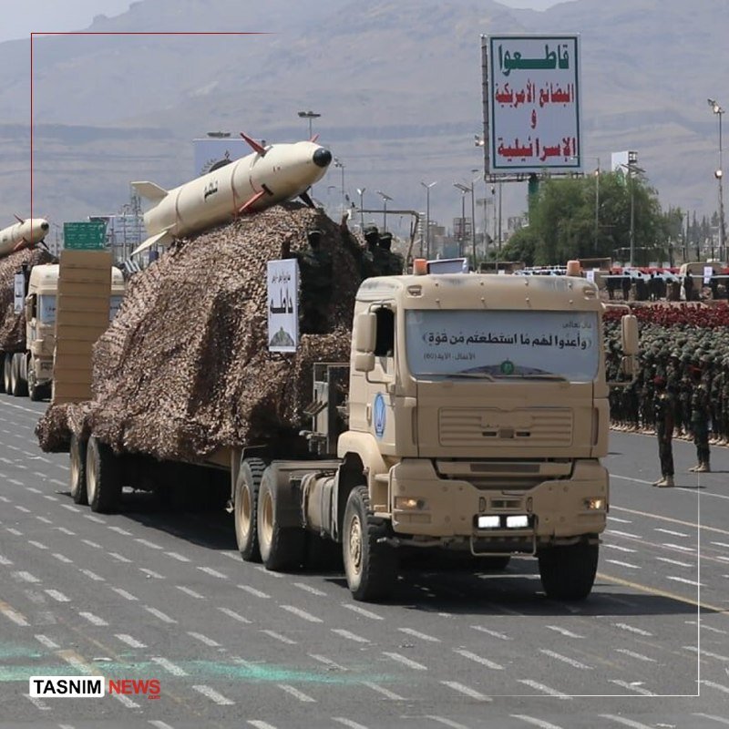 A camouflaged military truck carrying a large missile on its flatbed drives down a street lined with uniformed personnel and Arabic signage.
