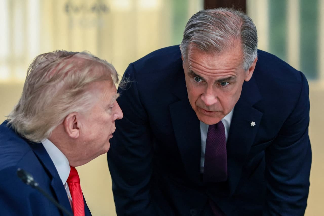Two men in dark suits, one seated with blonde hair and a red tie and the other leaning forward with a purple tie, appear to be conversing in a formal indoor setting.