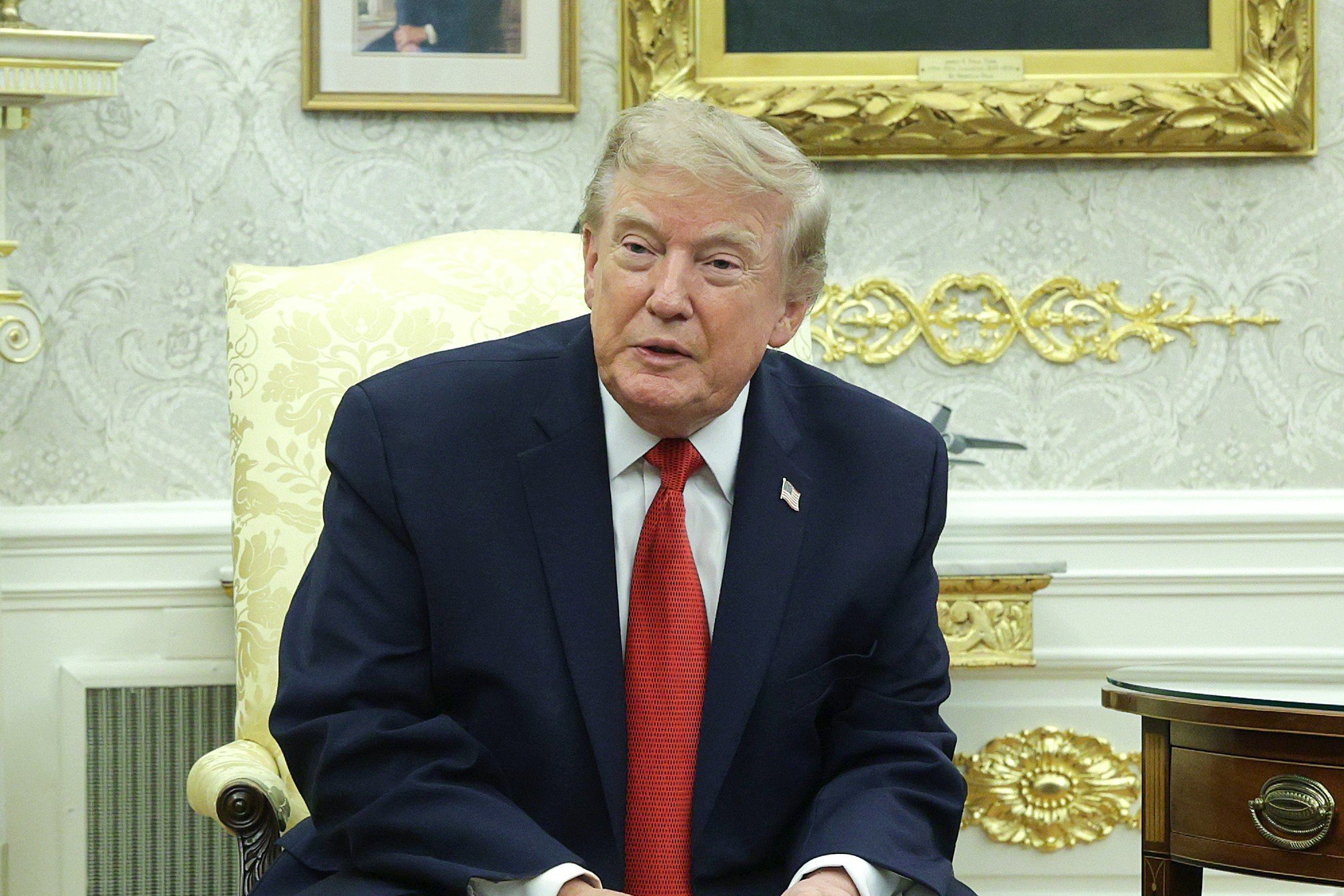 A blonde-haired man in a dark suit, white shirt, and red tie sits in a cream-colored upholstered chair in an ornately decorated room with gilded frames and patterned wallpaper.