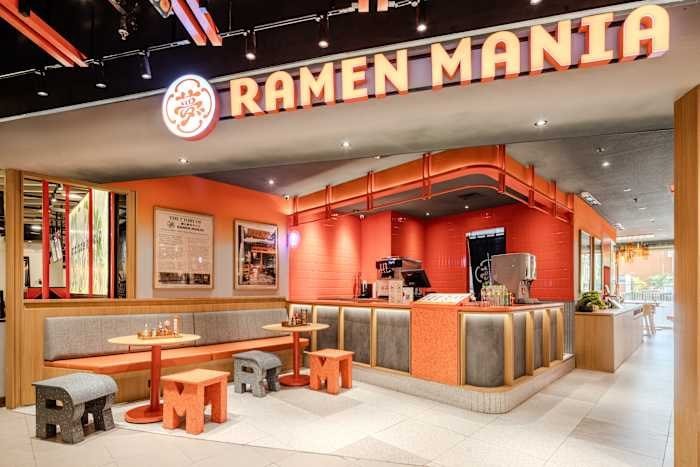 An empty orange-themed ramen restaurant interior features a service counter, booth seating, and a large illuminated "RAMEN MANIA" sign above the entrance.