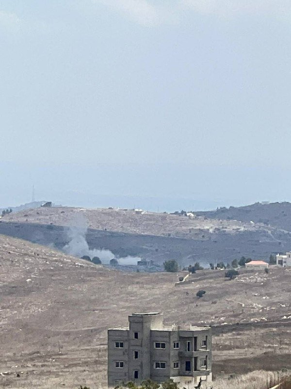 A plume of white smoke rises from an arid, hilly landscape behind an unfinished concrete building, with scattered structures visible in the distance under a hazy sky.
